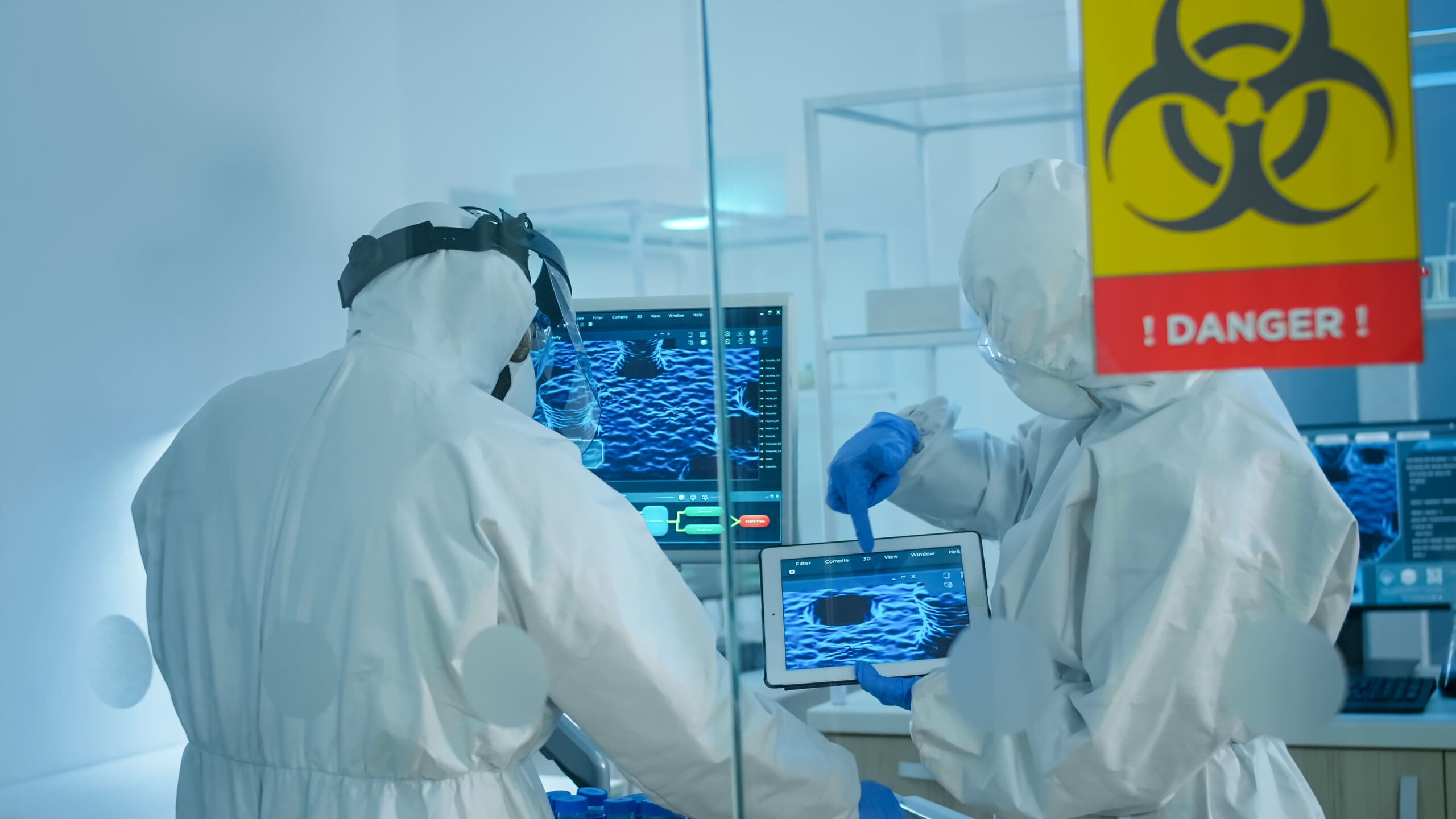 Scientists in coverall standing behind the glass wall working in danger area of lab. Chemists trying to develop vaccine using tablet examining virus evolution researching for treatment against covid19