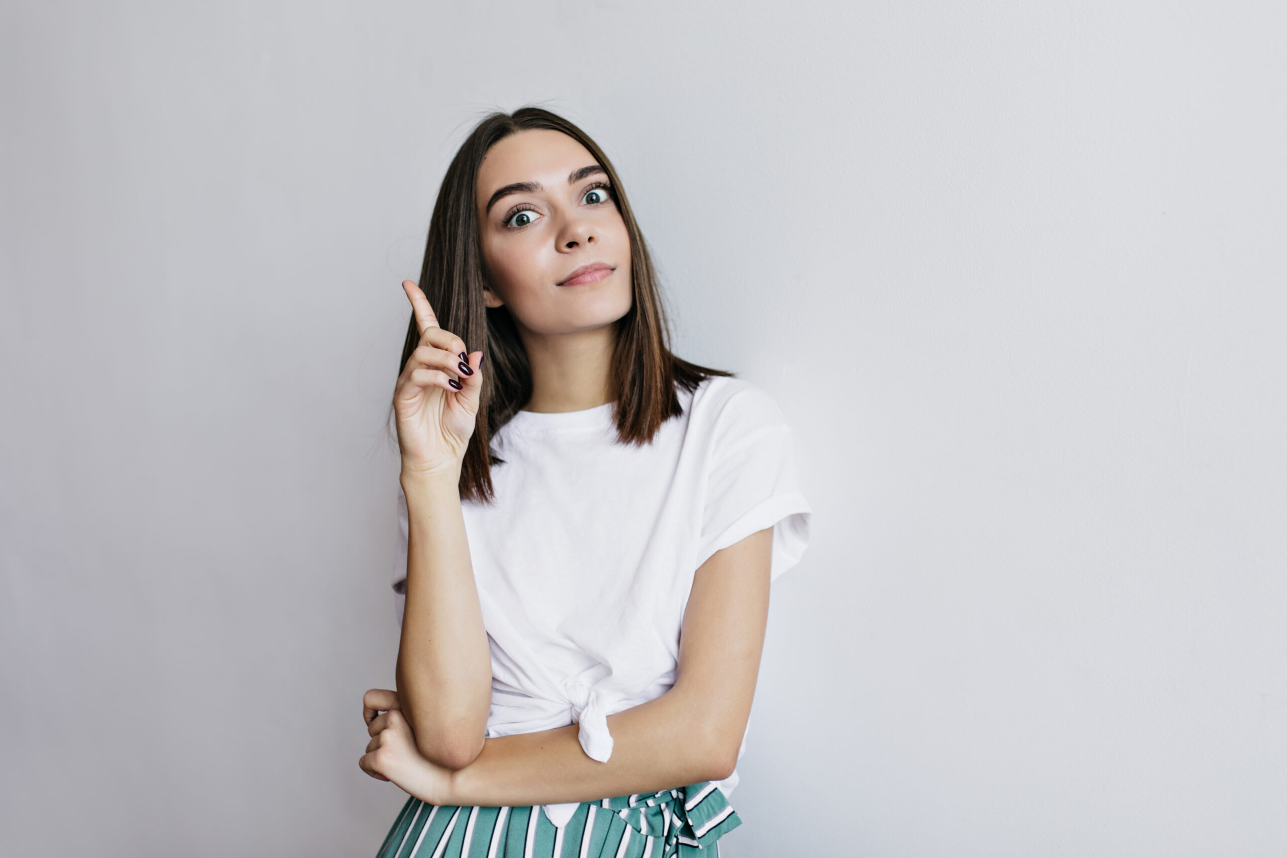 Joyful lady with dark eyes funny posing in studio. Female model in white t-shirt making faces during photoshoot.