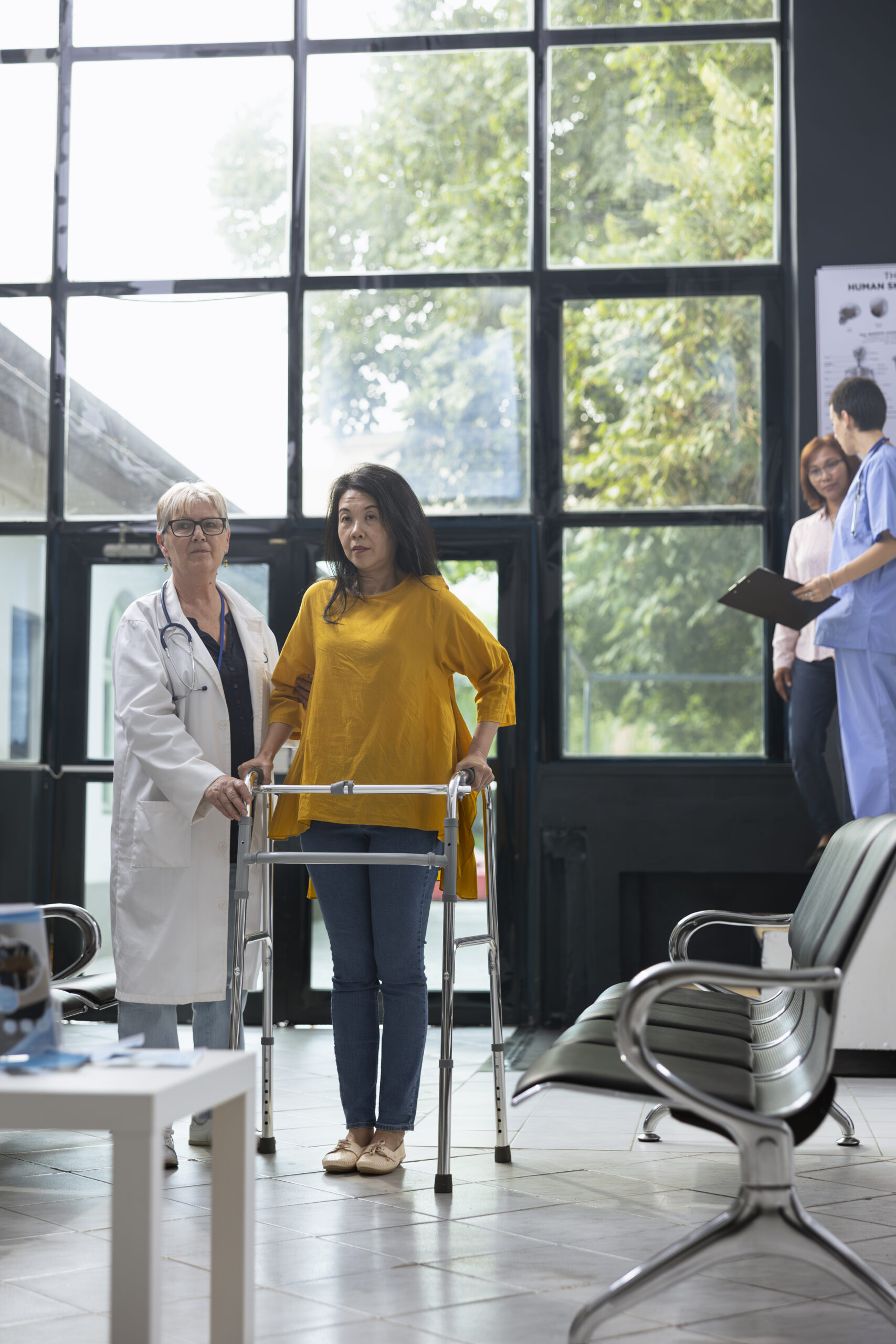 Asian woman using walking frame during rehabilitation healing process, receiving physical support from medical specialist at a health clinic. Moving around for recovery journey.