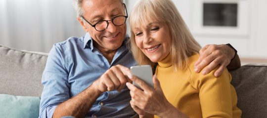 Happy Senior Couple Using Mobile Phone Together, Husband Pointing Finger At Cellphone Showing Something Browsing Internet Sitting On Couch At Home. Mature People And Gadgets